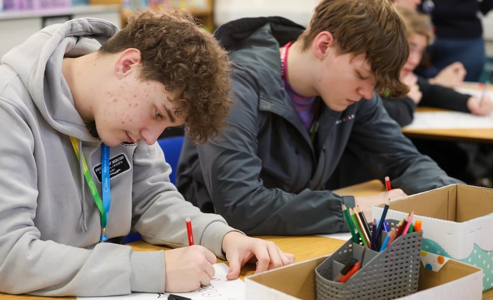 Two teenage boys seated at desks draw on paper with colored pencils; boxes of art supplies are on the desks in a classroom setting.