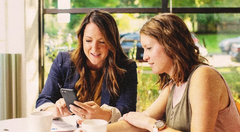 Two women sit at a table with coffee cups, looking at a smartphone together, in front of large windows with greenery outside.