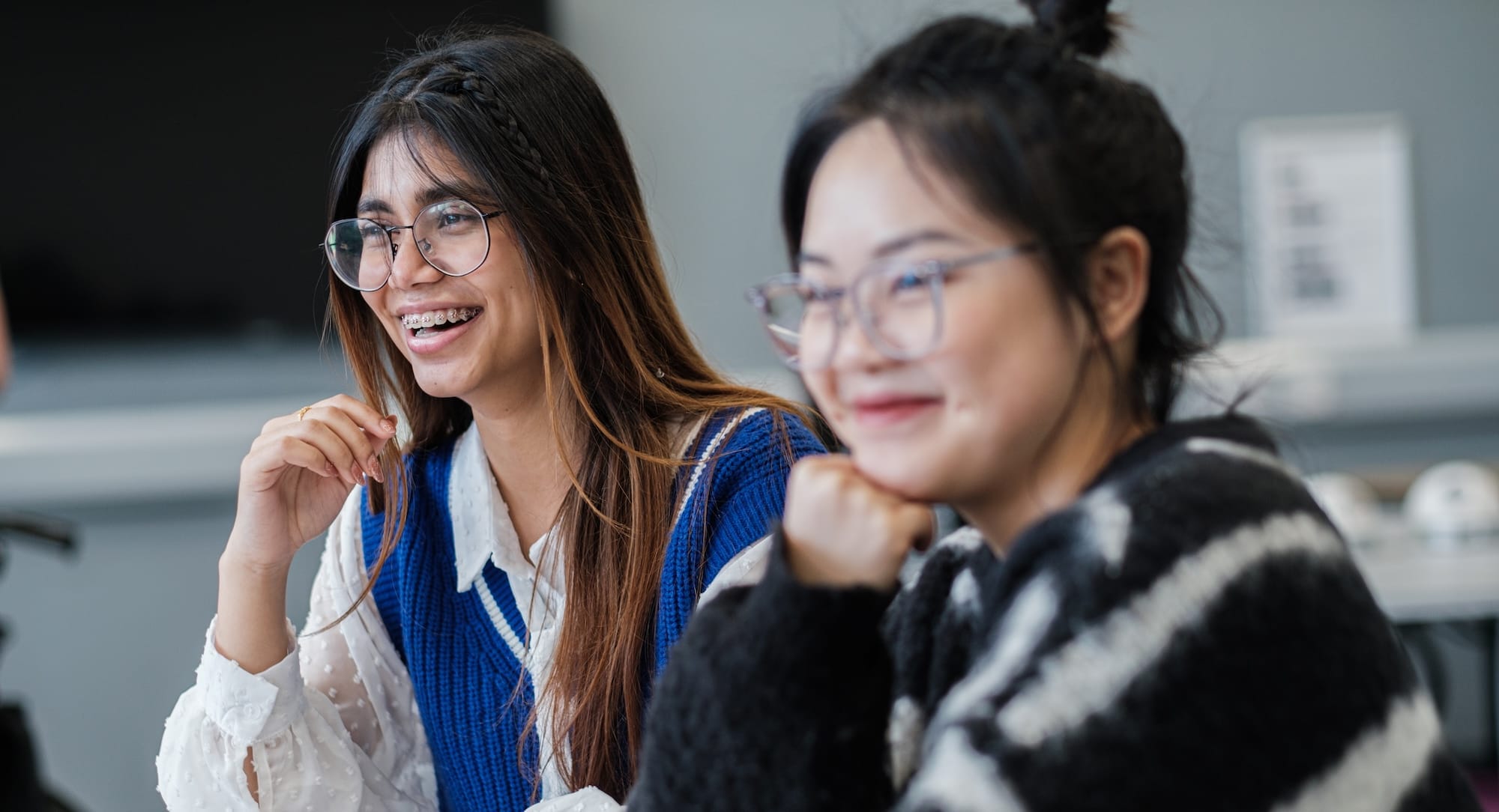 Two young women wearing glasses sit side by side, smiling and looking towards something off camera in a bright indoor setting.